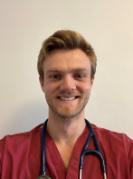 A man with short blond hair and a beard wearing maroon medical scrubs and wearing a stethoscope